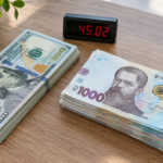 Долари й гривні поруч на столі з цифровим тікером що показує 45,02